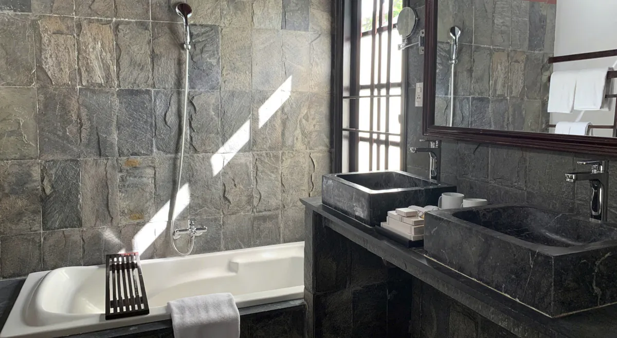 A slate-tiled bathroom: a bath with a shower, two dark stone basins and a wood-framed mirror