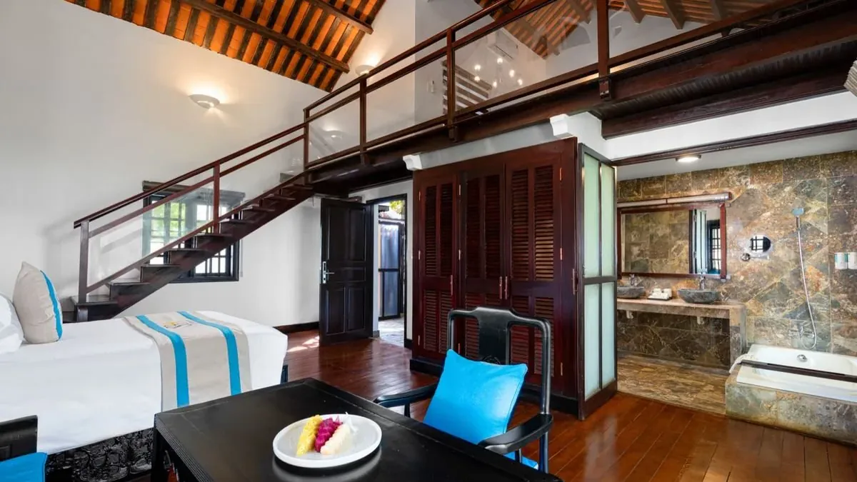 The ground floor of a duplex villa: the bed, the wooden stair to the mezzanine under the tiled roof and the stone bathroom to the right