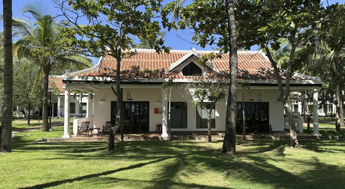 La fachada de una villa de planta baja entre los árboles del jardín, con su porche de columnas y el tejado de teja