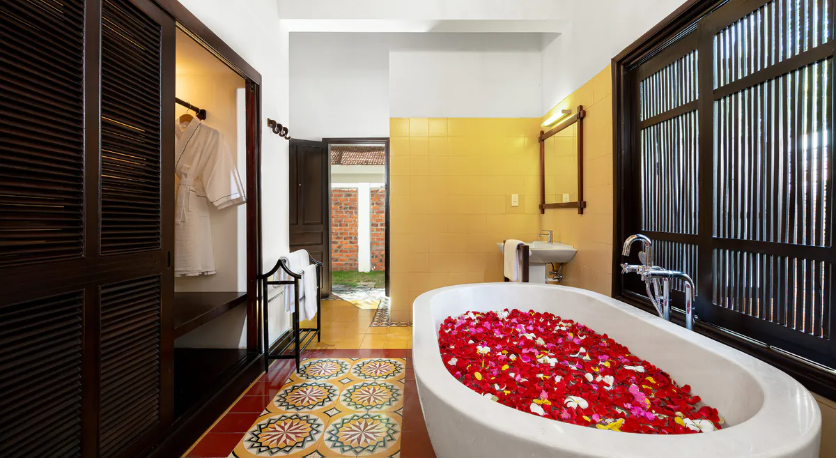 A villa bathroom at Le Belhamy: a freestanding bath filled with petals, yellow tiling, louvred shutters and an encaustic tile floor