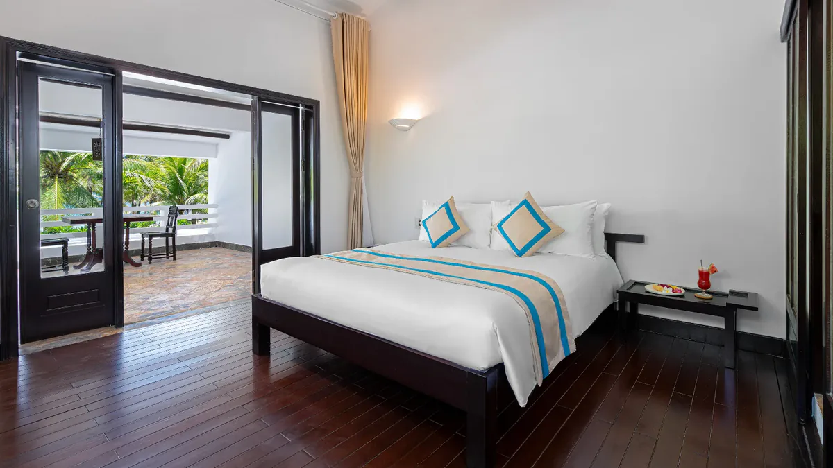 A bedroom opening through two sliding panels onto a terrace with a table and chairs, among the palms