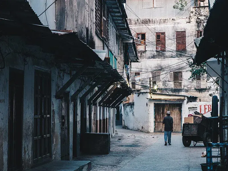 A Stone Town alley with a wooden arcade, overhead wiring and a man walking away with a cart
