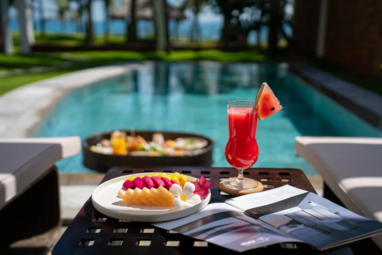 Un plato de fruta tallada y una copa de zumo de sandía junto a la piscina privada de una villa del Belhamy