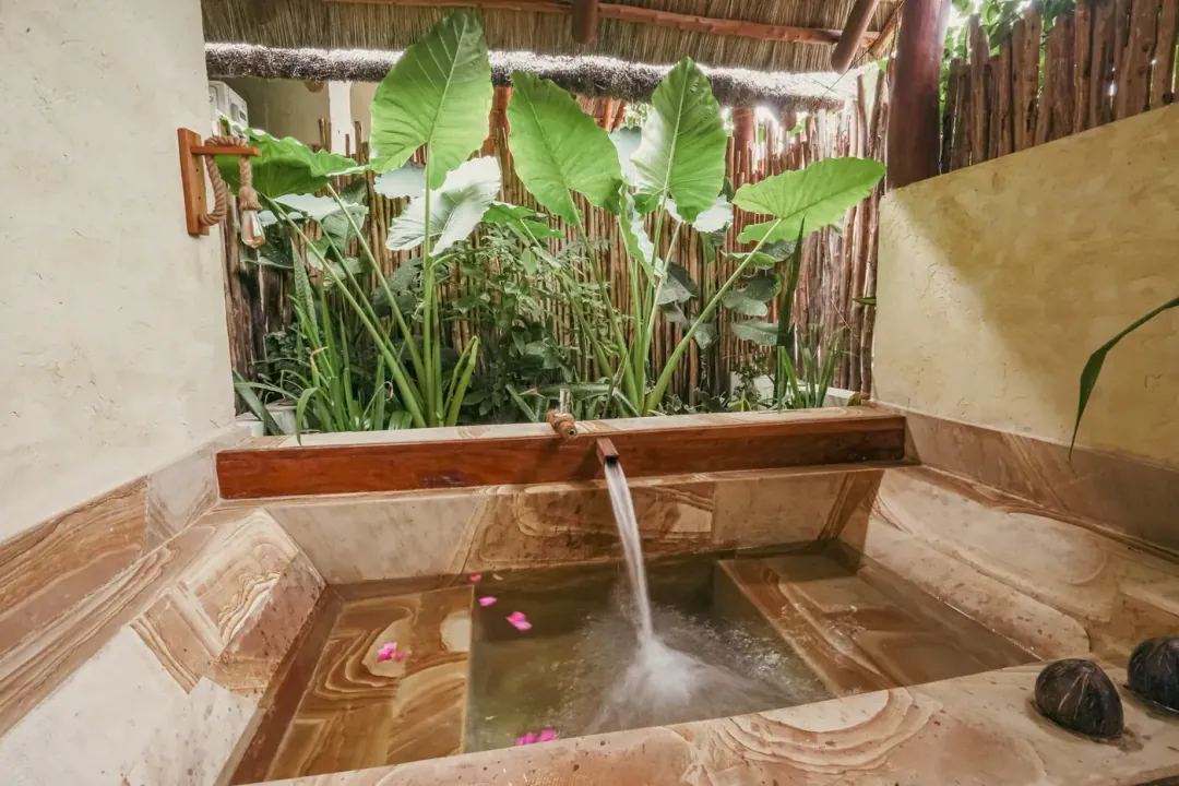 Bañera exterior de piedra con agua cayendo, entre plantas tropicales y cañas