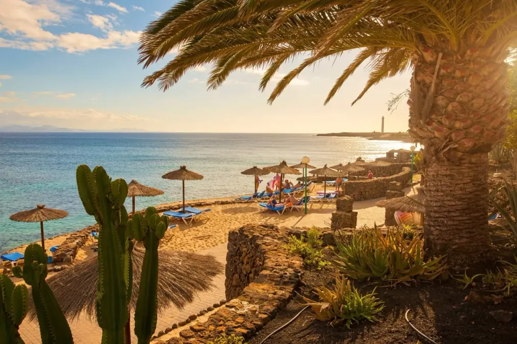 The beach at the Royal Mónica backlit at sunset: thatched parasols, sun loungers and the lighthouse beyond