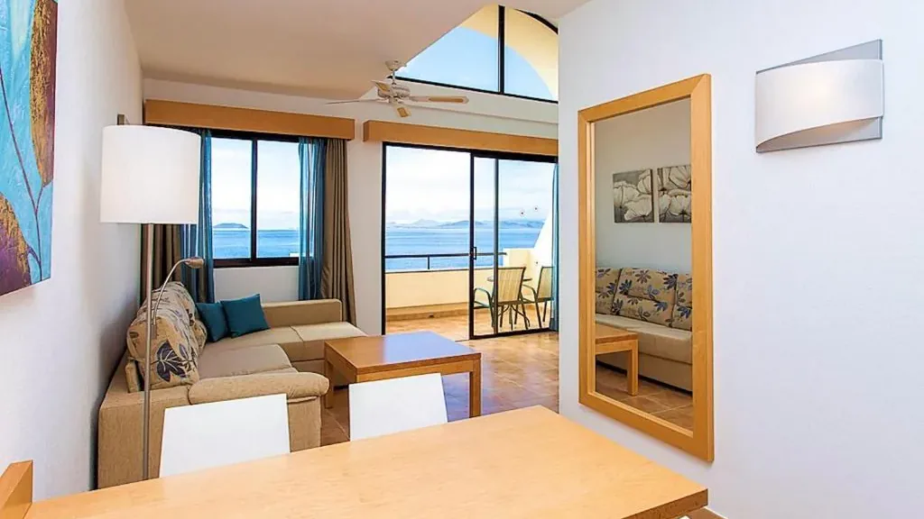 The living and dining area of a room at the Royal Mónica: sofa, table, a beech mirror and windows onto the sea