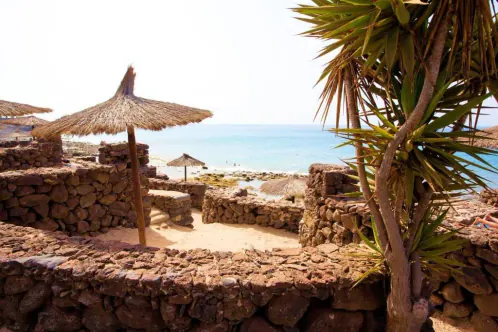 A pale-sand beach seen through a volcanic stone wall, with a thatched parasol