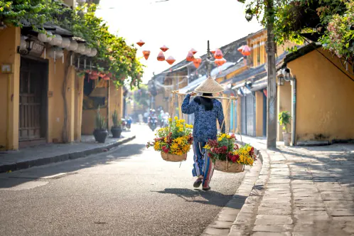 A street in Hội An's old town with ochre walls and a flower seller carrying her shoulder pole