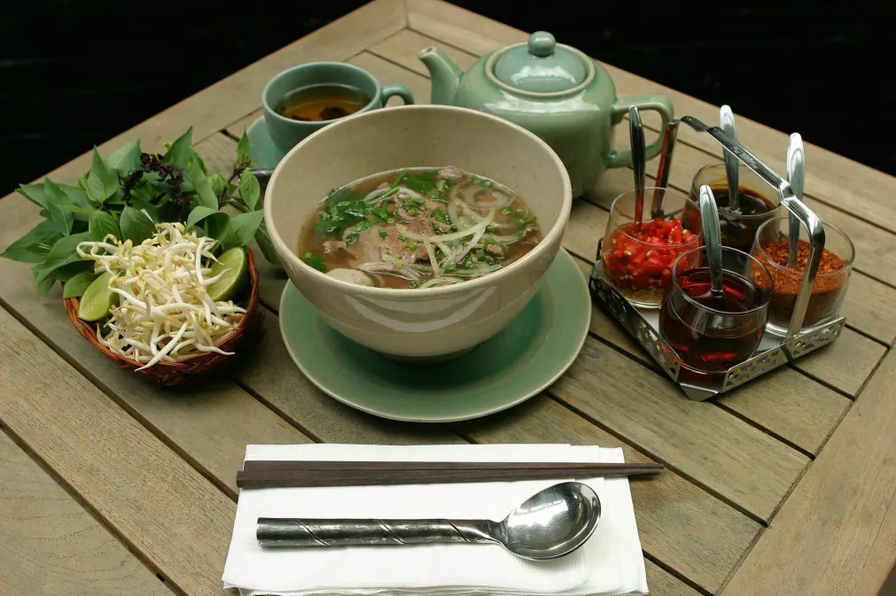 A phở served with its plate of herbs, sprouts and lime, on a teak table