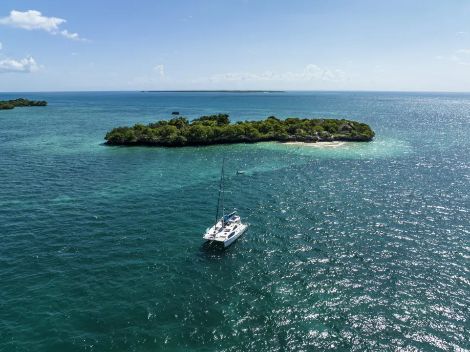Aerial view of a wooded islet with a spit of white sand and a catamaran anchored in turquoise water